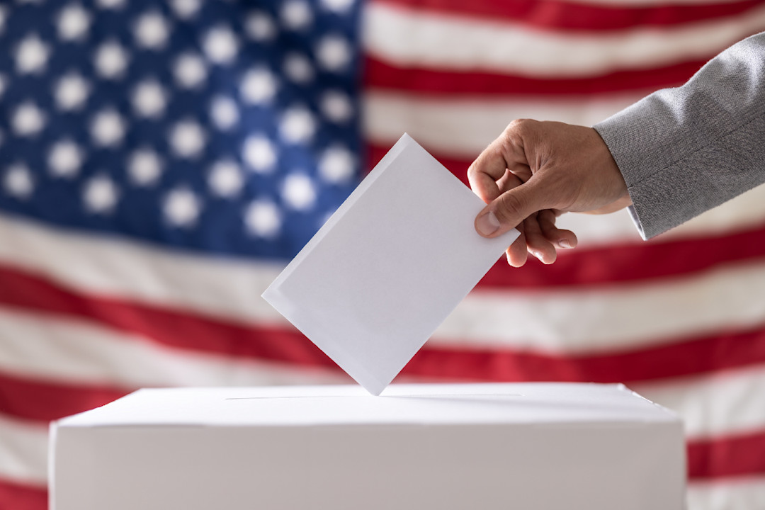 Hand holding ballot in voting ballot box with USA flag in background- GettyImages