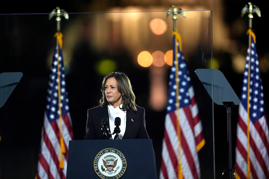 Kamala Harris standing giving a speech in from of US flags 	